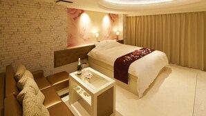 A hotel room with a large bed, a small side table with a bottle and glasses, a sofa with patterned cushions, and a brick wall. - Hotel venus NEO - Adults Only (Nagoya)