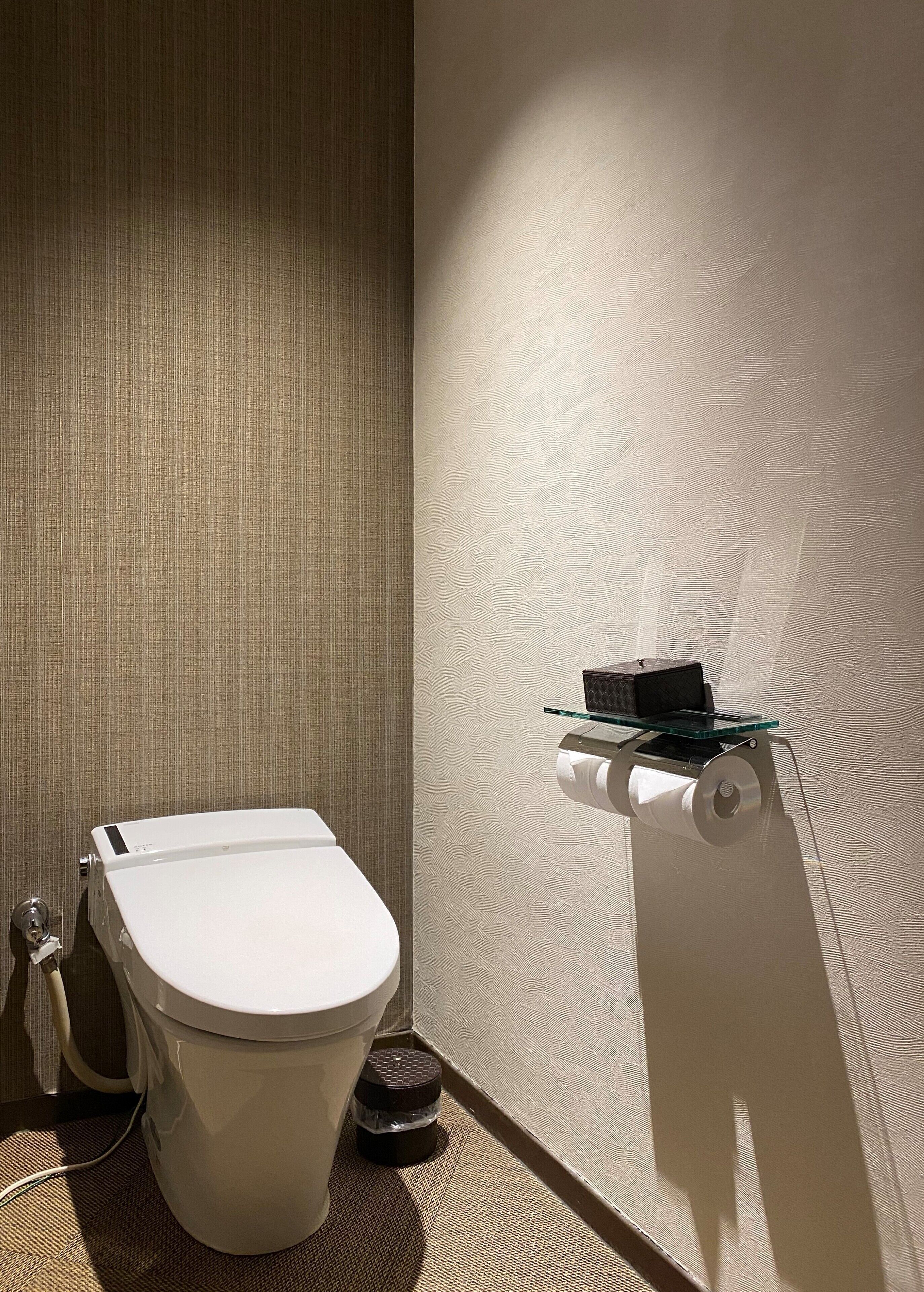 Eine Toilette mit weißem Deckel, ein kleines schwarzes Objekt auf einem Glastischchen und eine Toilettenpapierrolle an einem Halter.