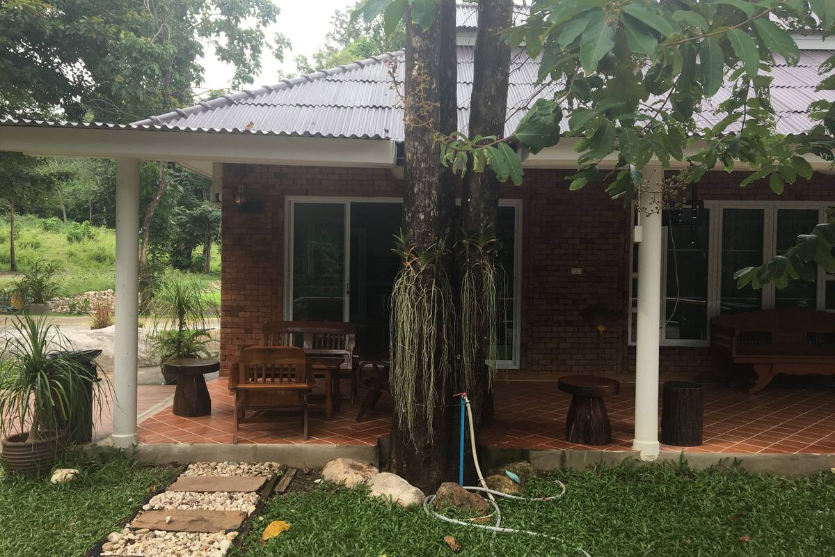 Een bakstenen huis met een overdekte patio, tuinmeubilair en een boom.