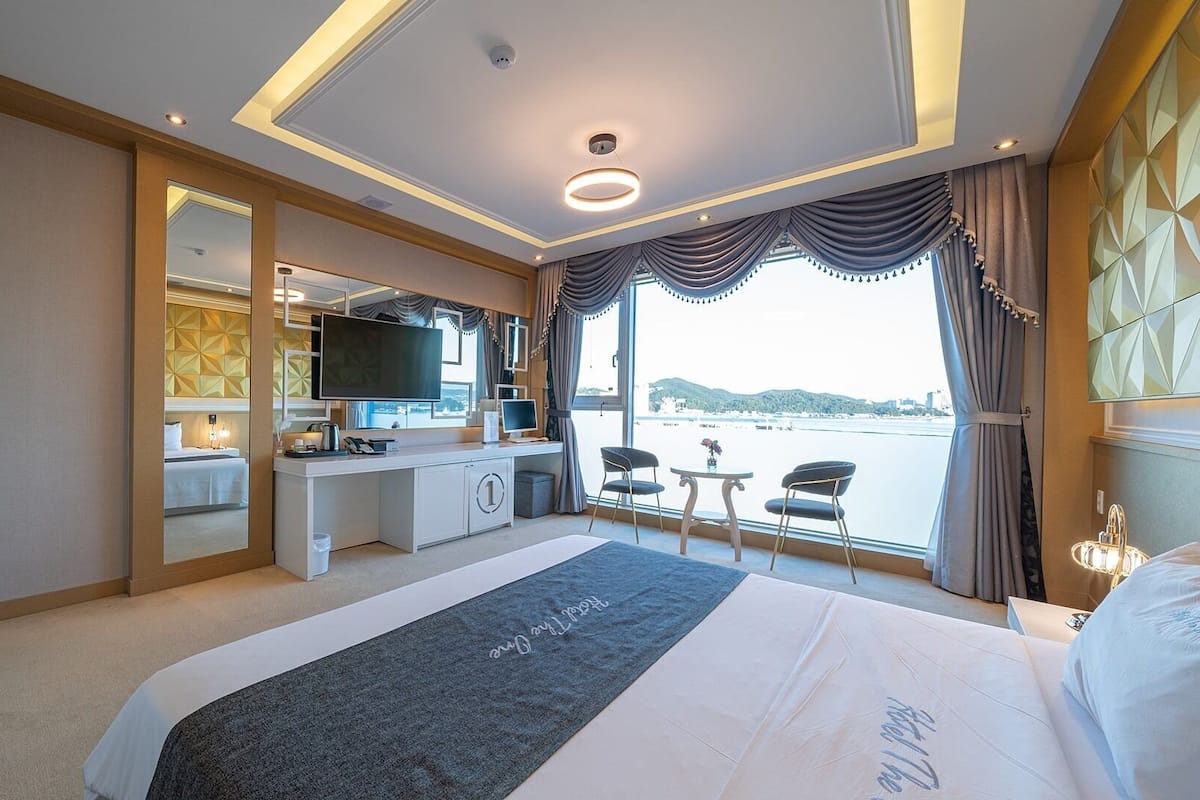 A hotel room with a bed, a desk, a TV, a view of the water, and a balcony.