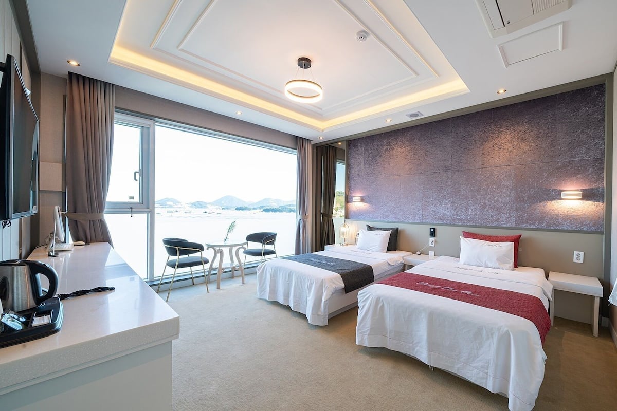 A hotel room with two beds, a desk with a kettle, a TV, and a view of the outdoors.