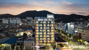 Un hotel de varios pisos llamado "Hotel The One" con una fachada moderna, situado en un paisaje urbano con edificios circundantes y una montaña al fondo.