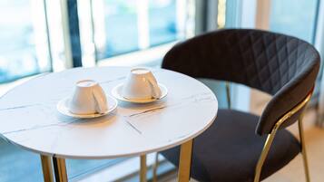 A round table with two white cups on saucers, a dark upholstered chair, and a view through large windows.