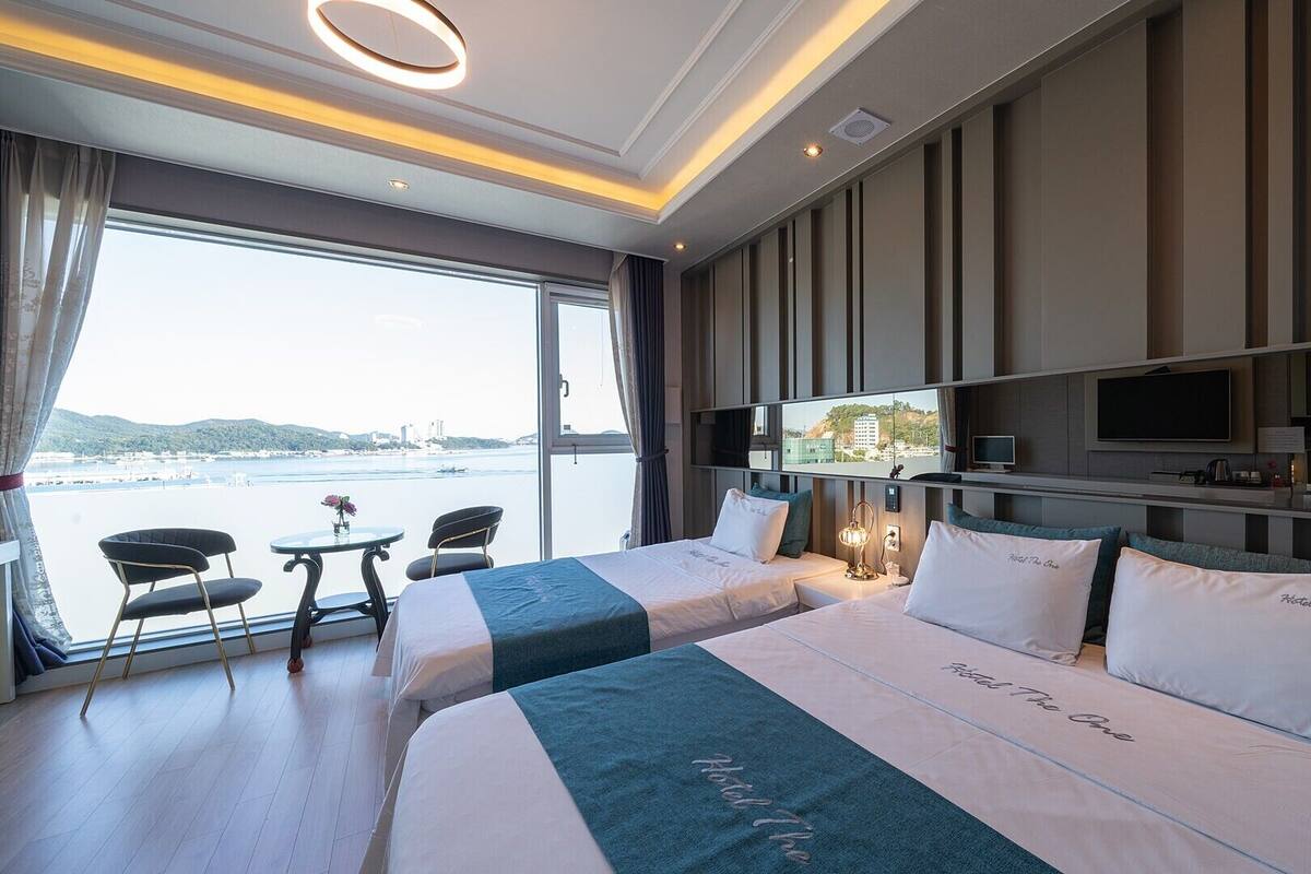 A hotel room with two beds, a TV, a desk, and a view of the sea.