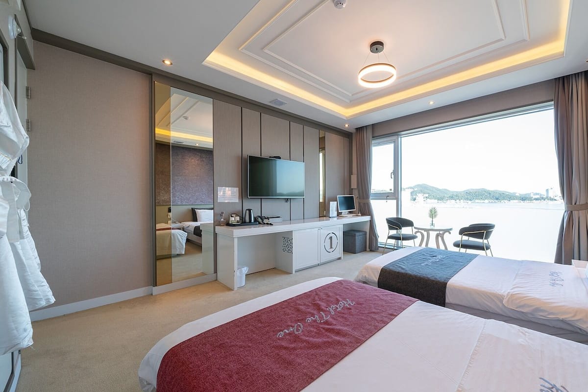 A hotel room with two beds, a desk with a computer, a TV, and a view of the sea.