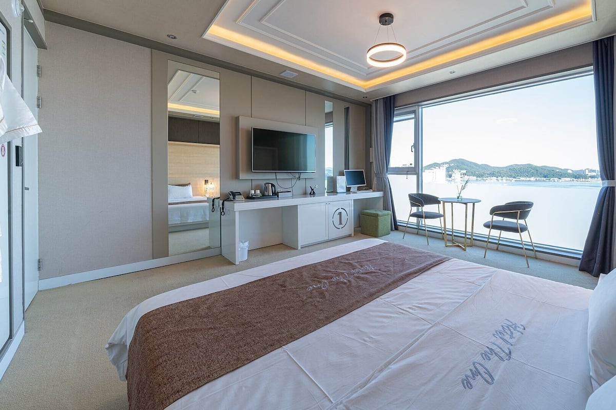 A hotel room with a large bed, a desk with a TV, a computer, and a view of the sea.
