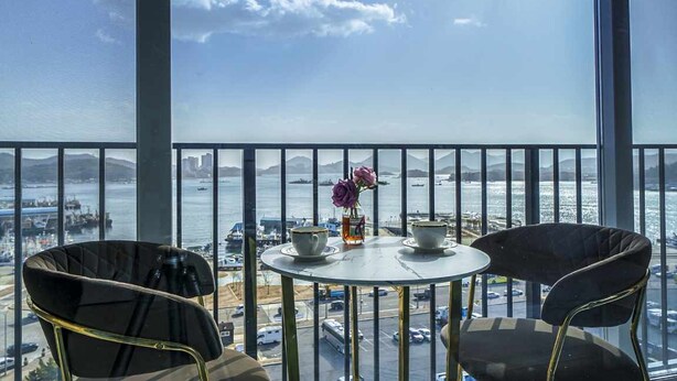 Un balcón con mesa y sillas, con vista a un puerto con barcos y un horizonte urbano distante.