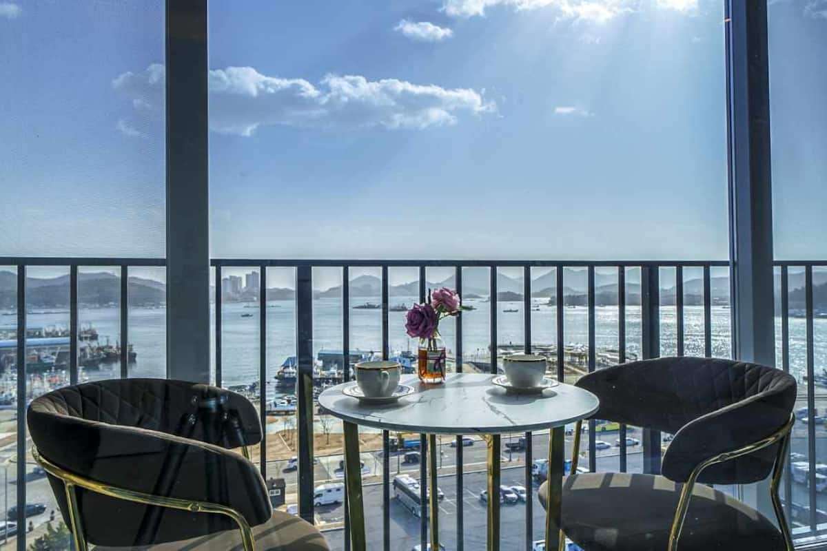 A balcony with a table and chairs, overlooking a harbor with boats and a distant cityscape.