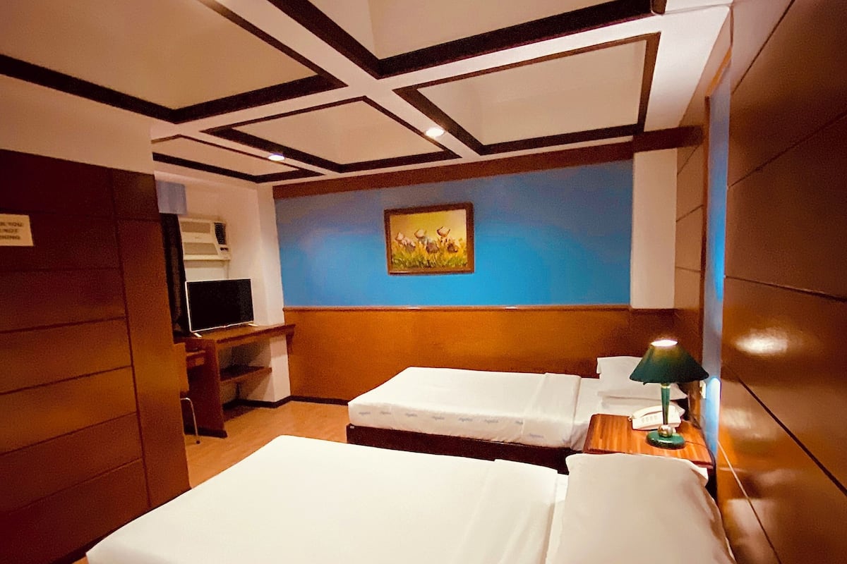 A hotel room with two beds, a desk, a chair, a painting on the wall, and a ceiling with a geometric design.
