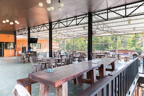An outdoor dining area with wooden tables and chairs, a covered roof structure, and a view of greenery in the background.