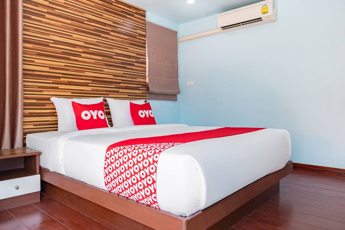 Une chambre d’hôtel moderne avec une tête de lit en bois, un lit avec une literie blanche et des touches de rouge, une table de chevet et un climatiseur mural.