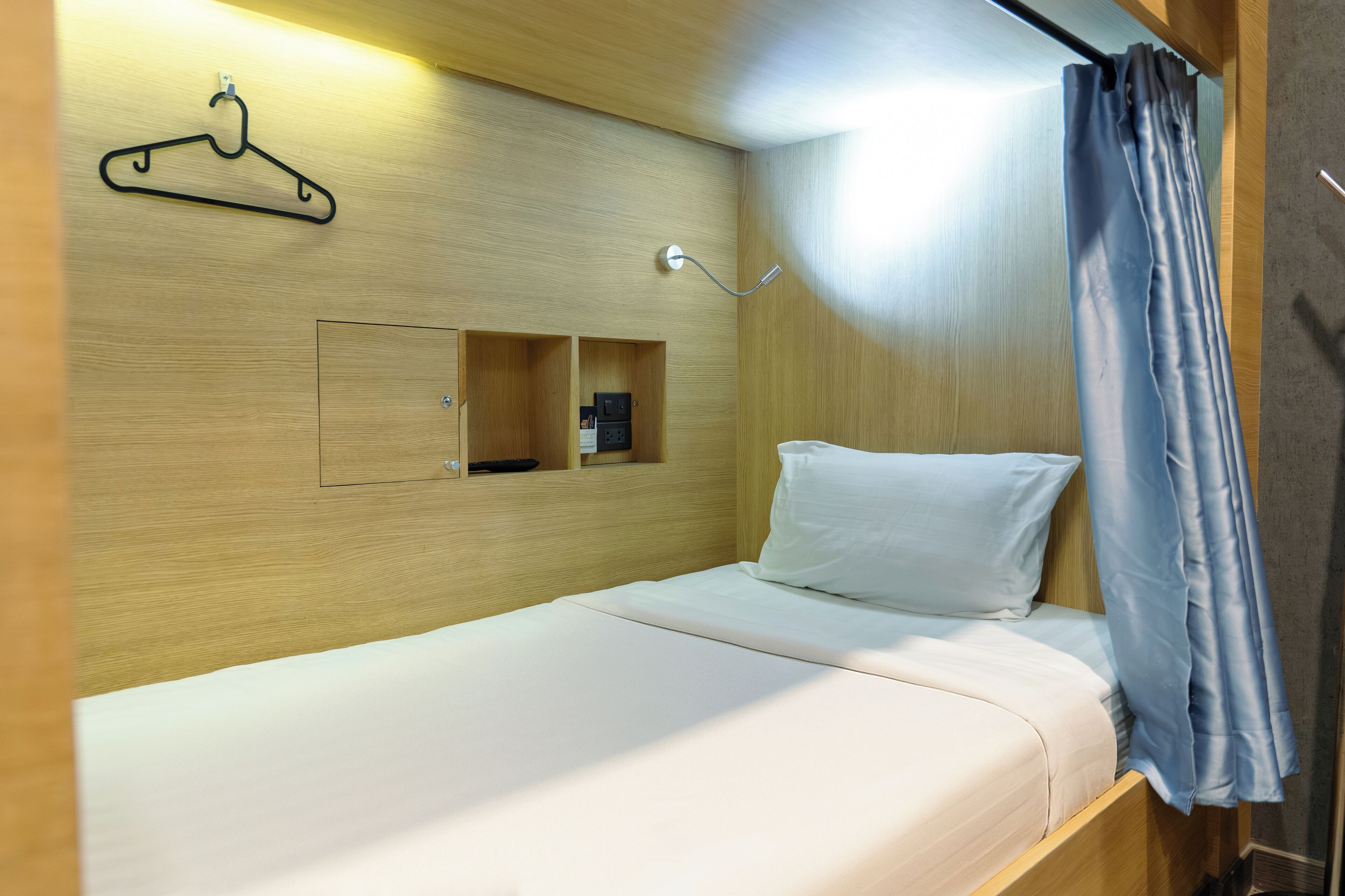 Una cama individual con colchón y almohada blancos, cabecera de madera, perchero y un estante con toma de corriente.