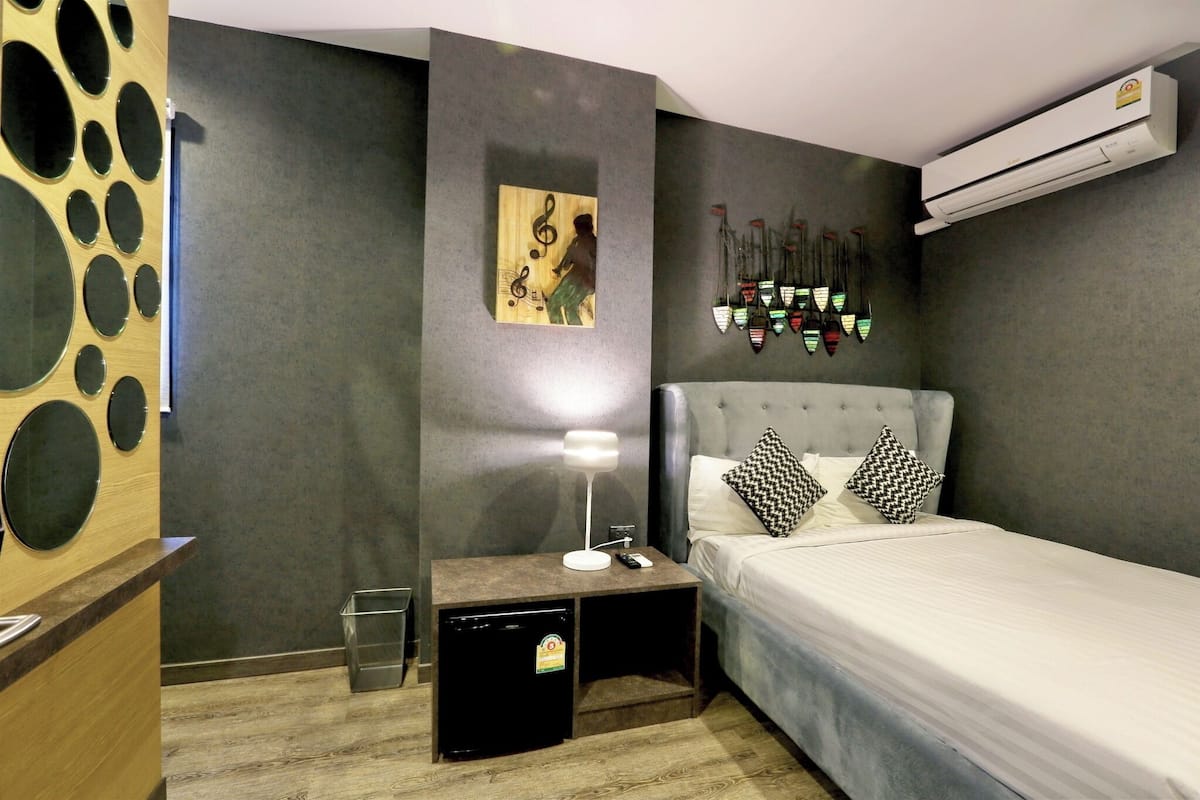 A hotel room with a grey bed, a small bedside table with a lamp, a wall-mounted air conditioner, and a decorative wall piece.