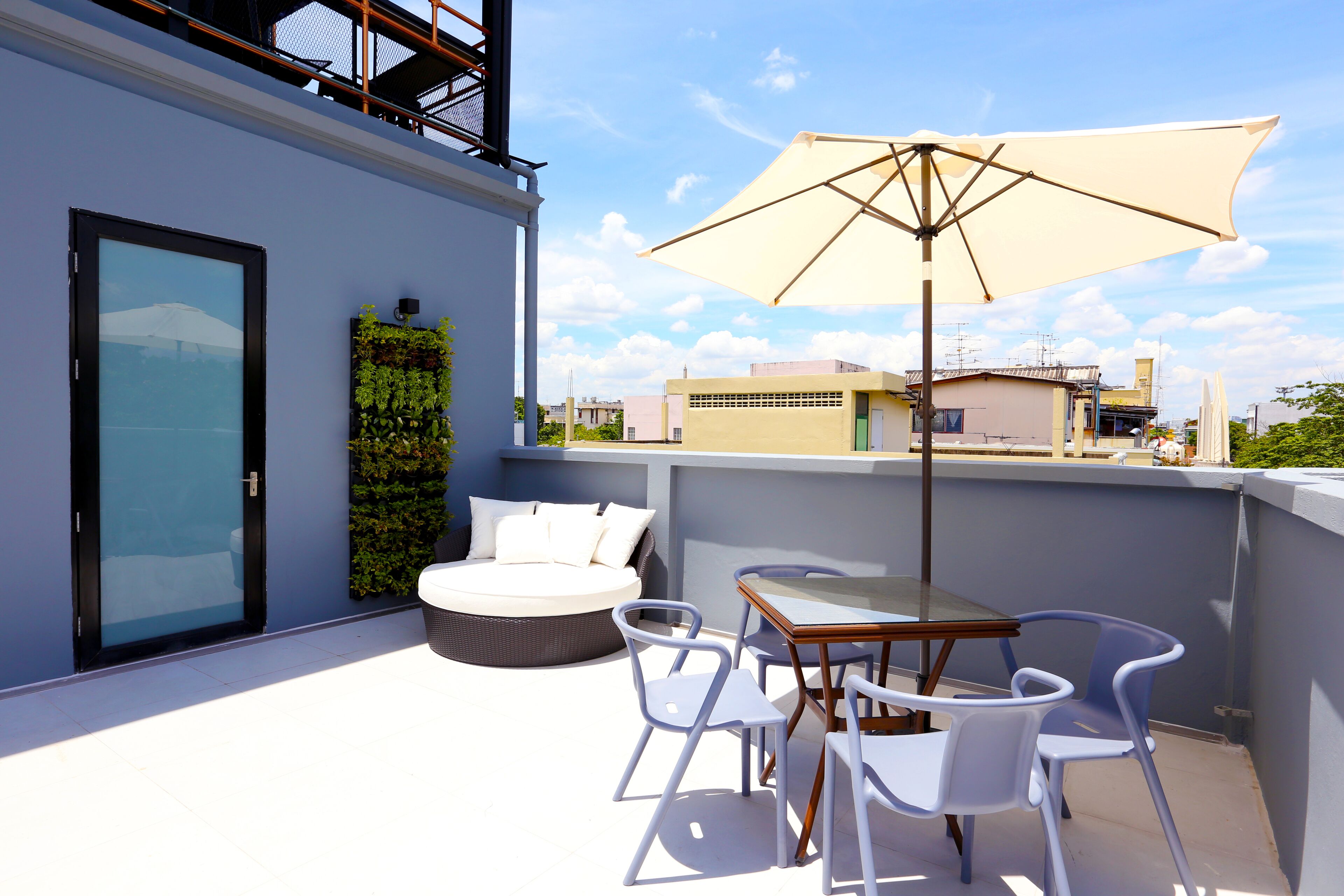 A rooftop terrace with a glass door, a round seating area with cushions, a table with chairs, and a large umbrella.