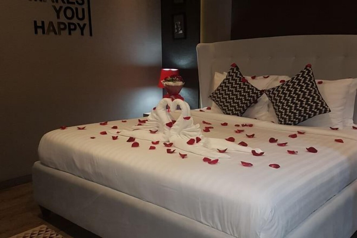 A neatly made bed with rose petals and a heart-shaped towel arrangement, white pillows, and a wall sign saying "MAKE YOU HAPPY".