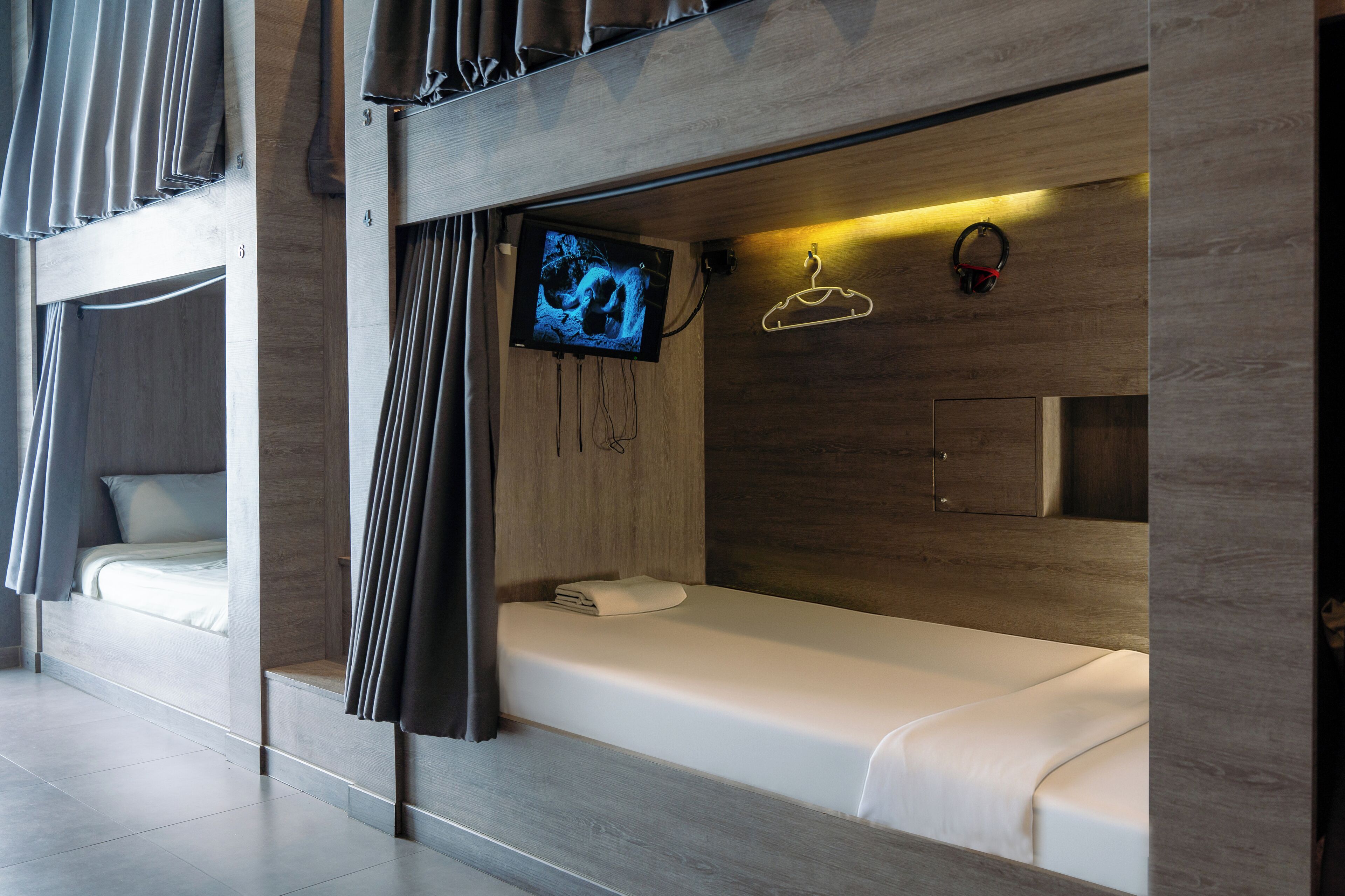 A modern hotel room with a bunk bed, a flat-screen TV mounted on the wall, a bedside table, and a hanging coat rack.