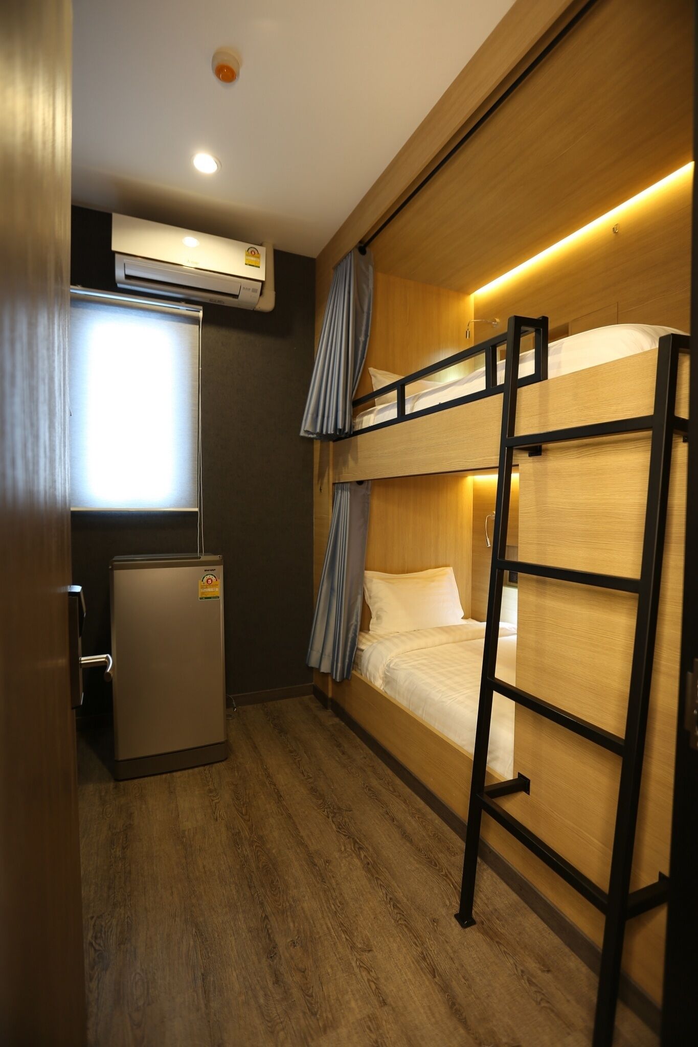 Habitación de hotel compacta con literas, un refrigerador pequeño y un equipo de aire acondicionado.