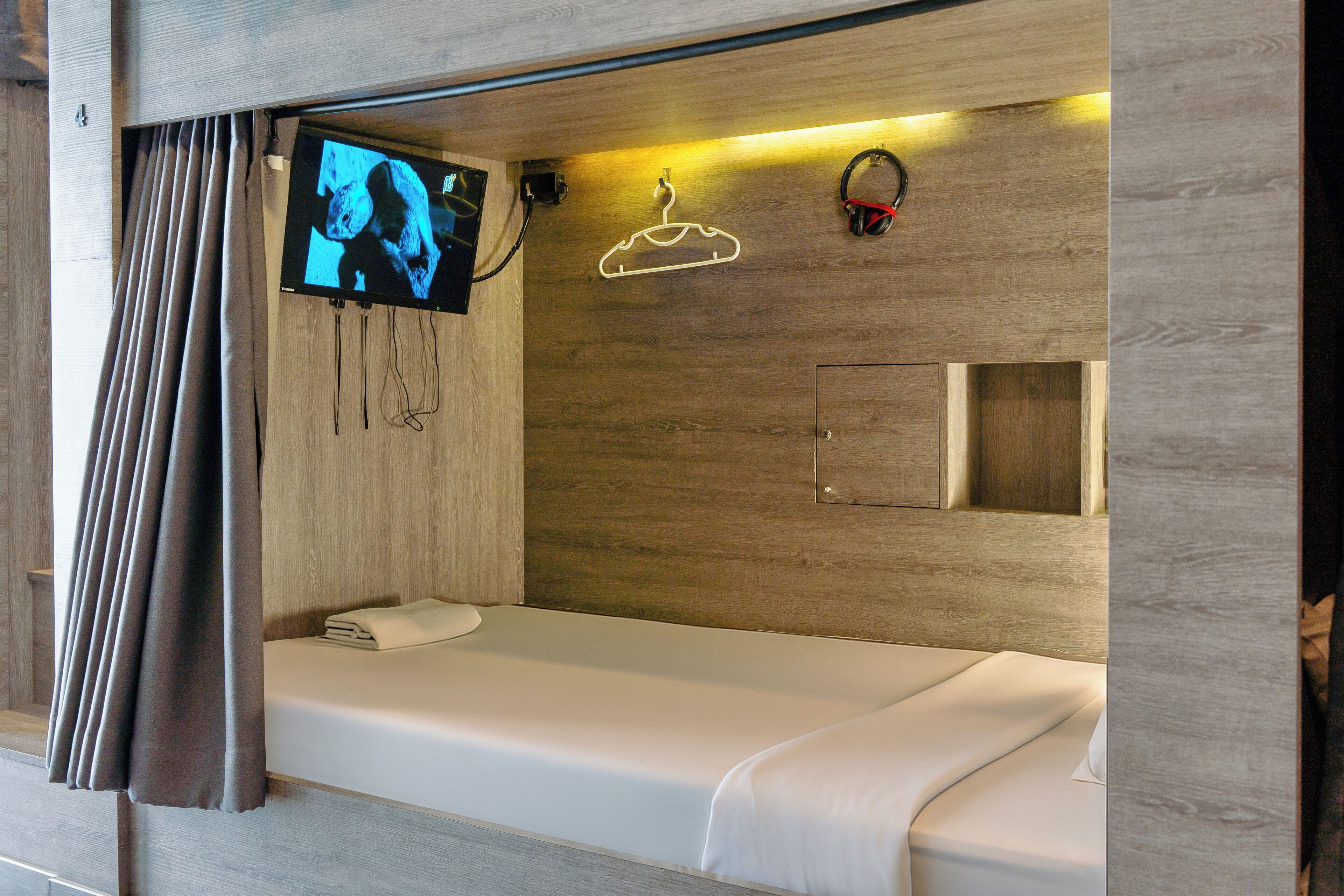 A single bed with a white mattress and a small bedside table, a television mounted on the wall, and a wooden headboard.