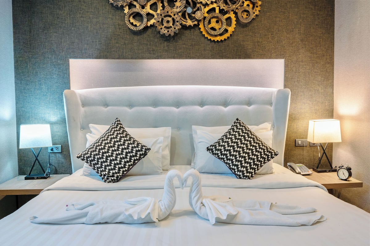 A hotel room with a tufted headboard, two patterned pillows, a bed with towels arranged in a heart shape, a bedside phone, and a wall clock.