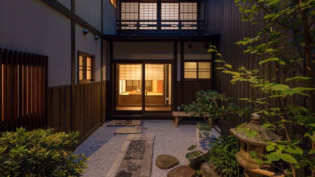 En traditionell japansk byggnad med en träfasad, en stensatt gång som leder till entrén och en liten trädgårdsdel med stenar och växter.