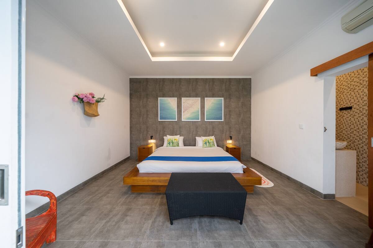 A modern bedroom with a large bed, bedside tables, a bench, and a wall-mounted flower arrangement.