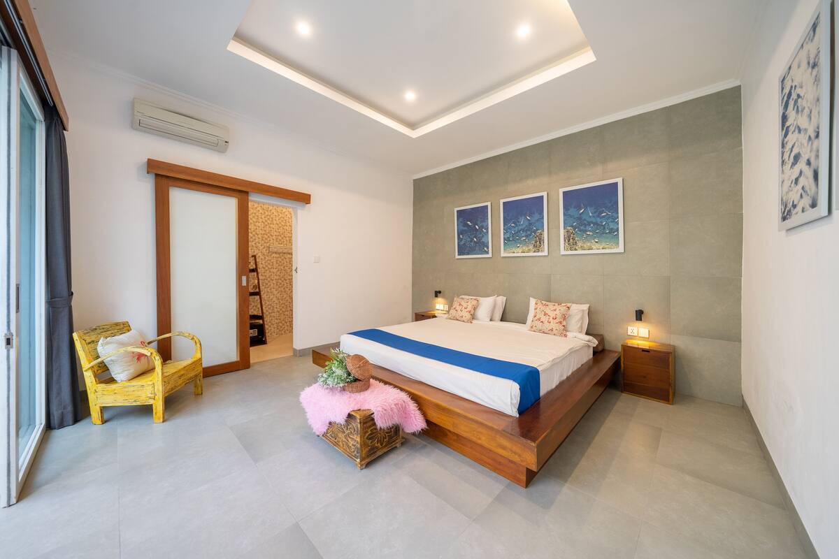A modern bedroom with a large bed, wooden headboard, and a small bedside table. There is a chair with a cushion and a decorative box on the floor.