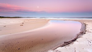Una playa con una superficie acuática lisa y reflectante y una luna creciente en el cielo.