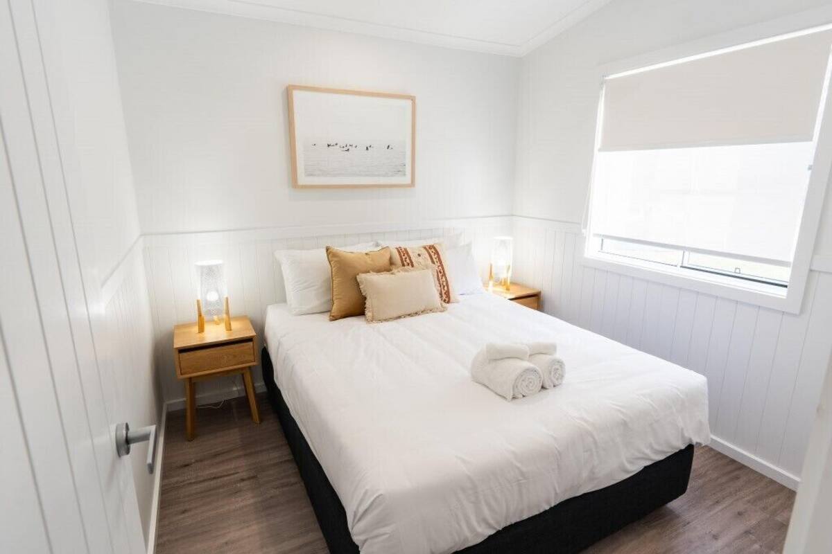 A neatly made bed with white linens, a framed picture on the wall, and a window with blinds.