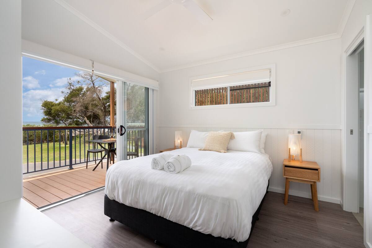 A bedroom with a large bed, a small table, a chair, and a balcony with a view of the outdoors.