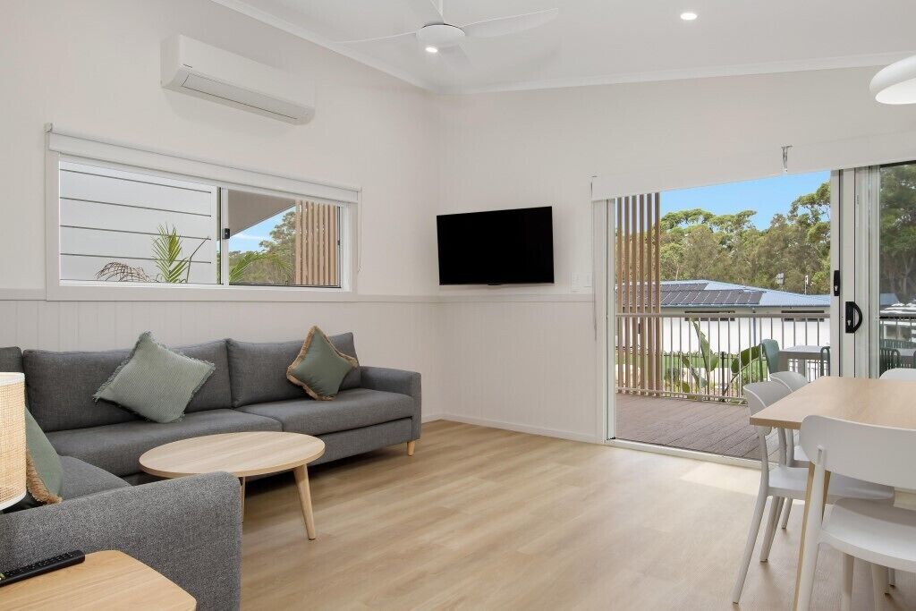 A modern living room with a grey sofa, a round coffee table, a flat-screen TV mounted on the wall, and a sliding glass door leading to an outdoor area.