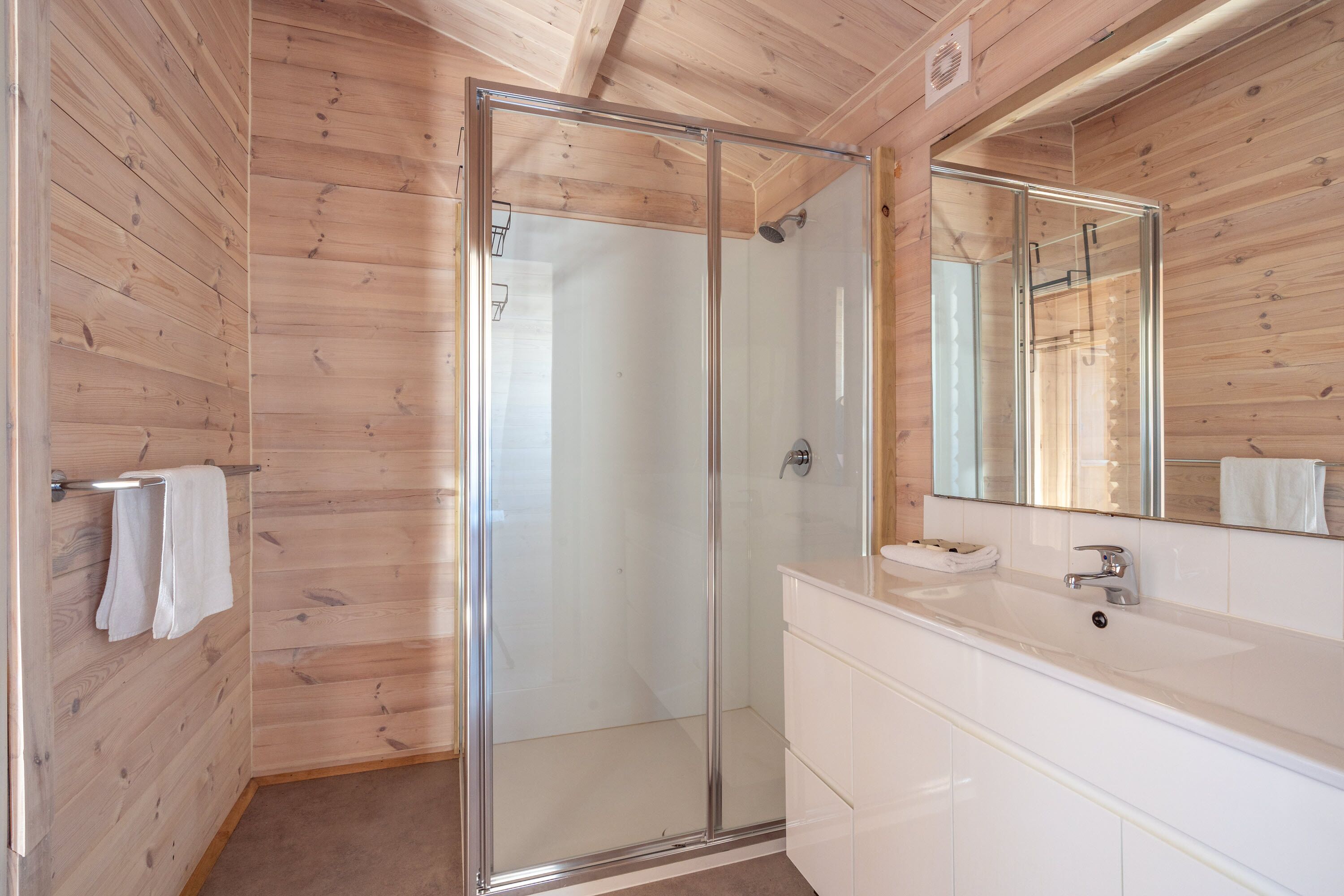 Un baño con paneles de madera, cabina de ducha de vidrio, lavamanos blanco y espejo.