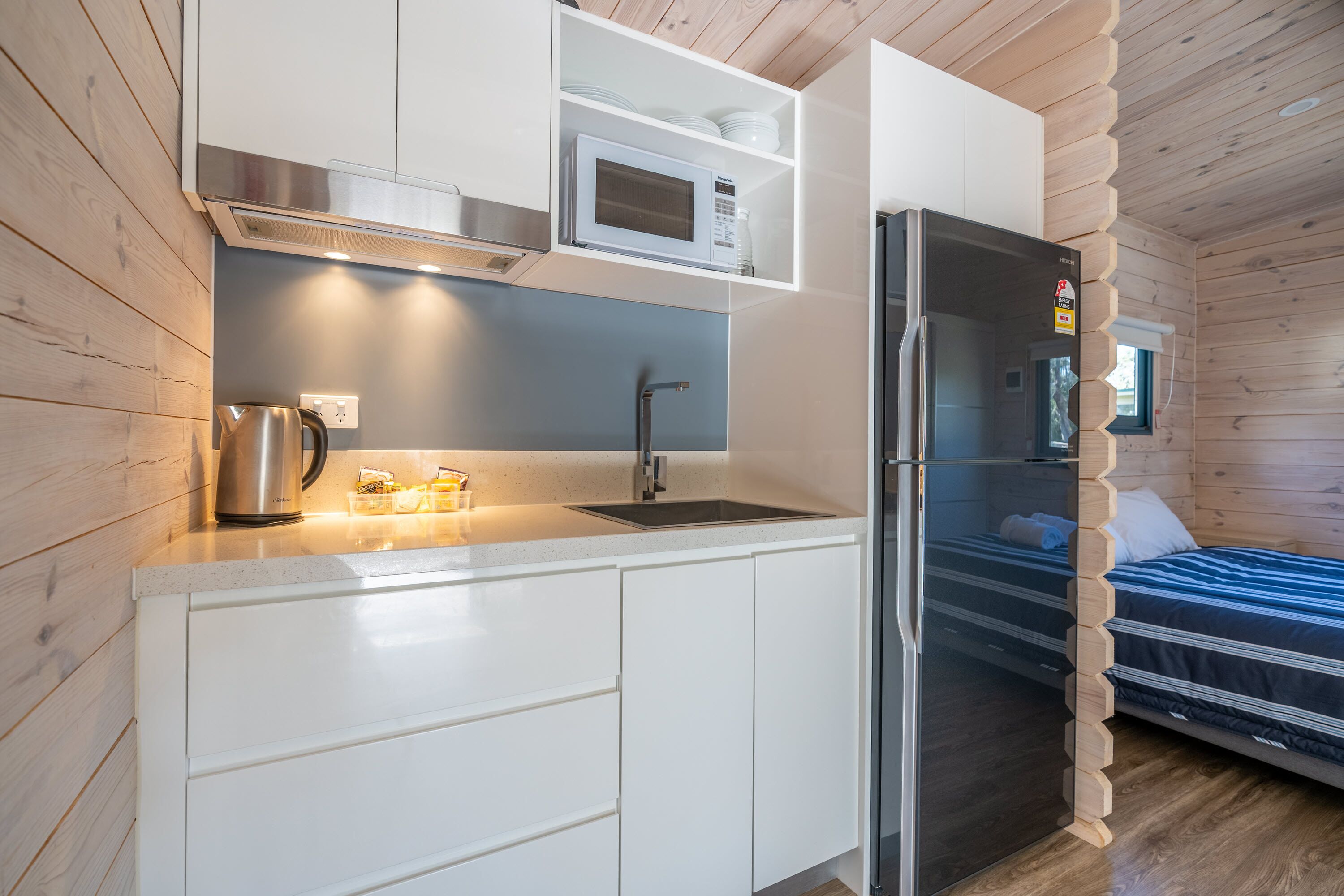 A compact kitchen with a microwave, sink, and stainless steel kettle. A bed with a wooden headboard is visible in the adjacent room.