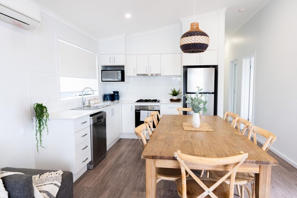 A modern kitchen with white cabinetry, a wooden dining table, and a hanging light fixture.