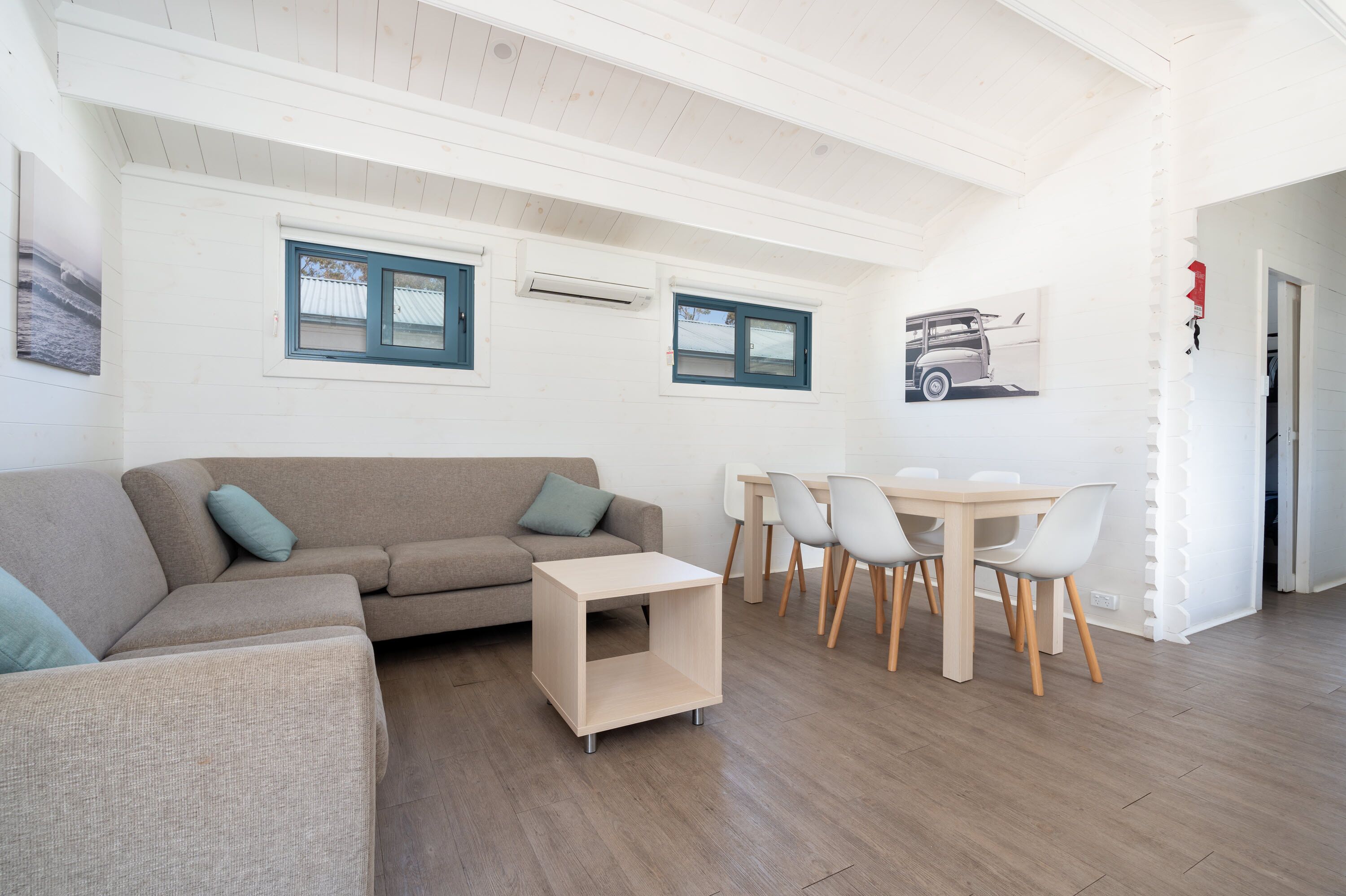 A modern living room with a sofa, a small table, and a dining area with chairs. There are two windows with blue frames and an air conditioning unit above them.