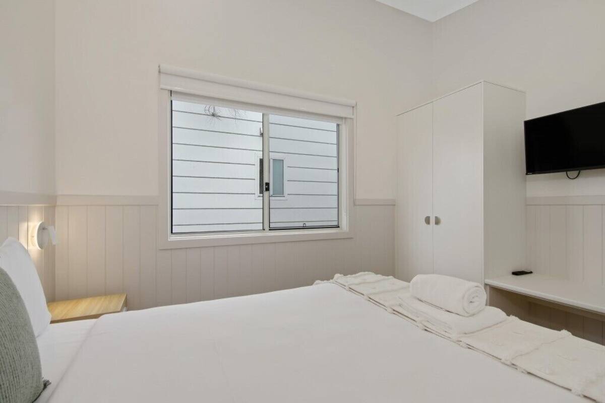 A bedroom with a bed, a window with blinds, a wardrobe, and a flat-screen TV mounted on the wall.