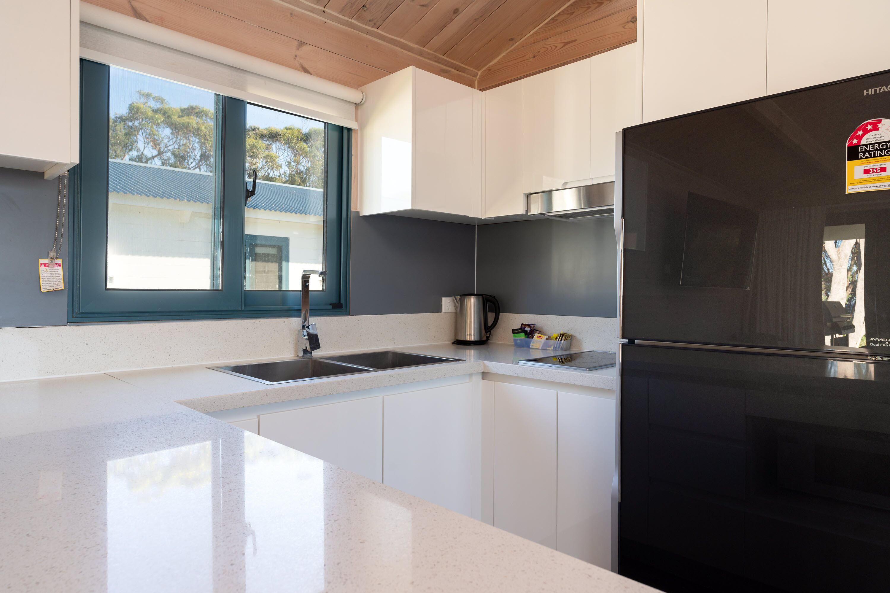 A modern kitchen with white cabinetry, a black refrigerator, a stainless steel sink, and a window with a view of trees.