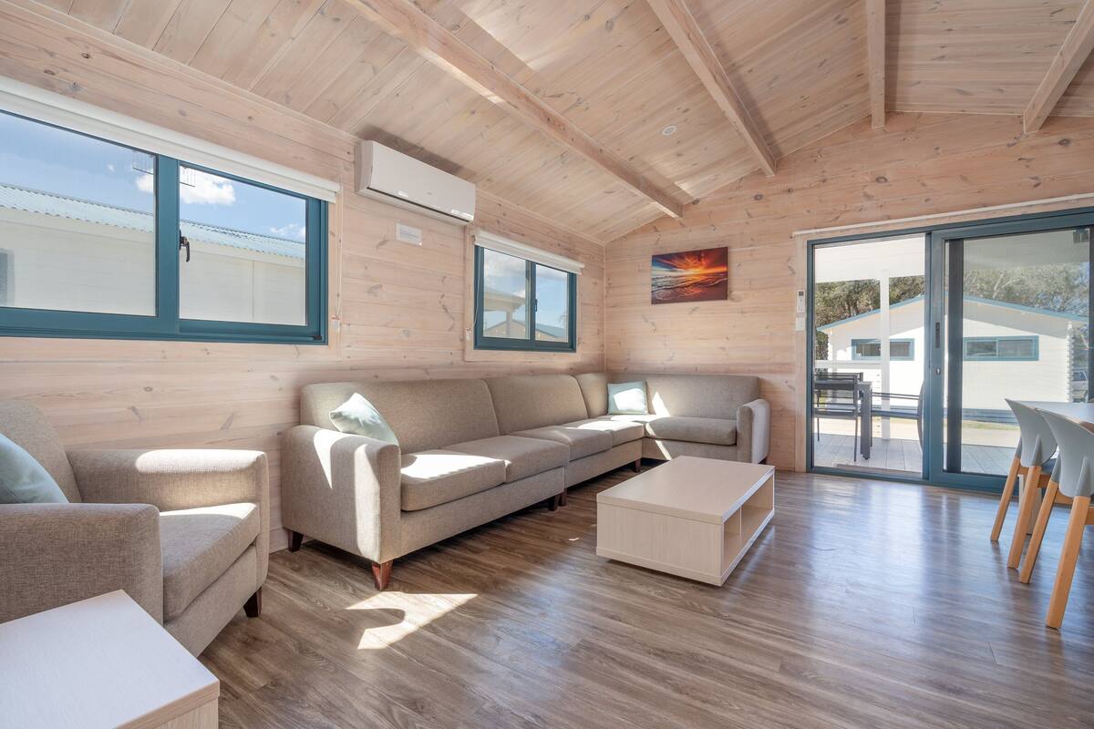 A cozy cabin interior with a wooden ceiling, a beige sofa, a small table, and a sliding glass door leading to an outdoor area.