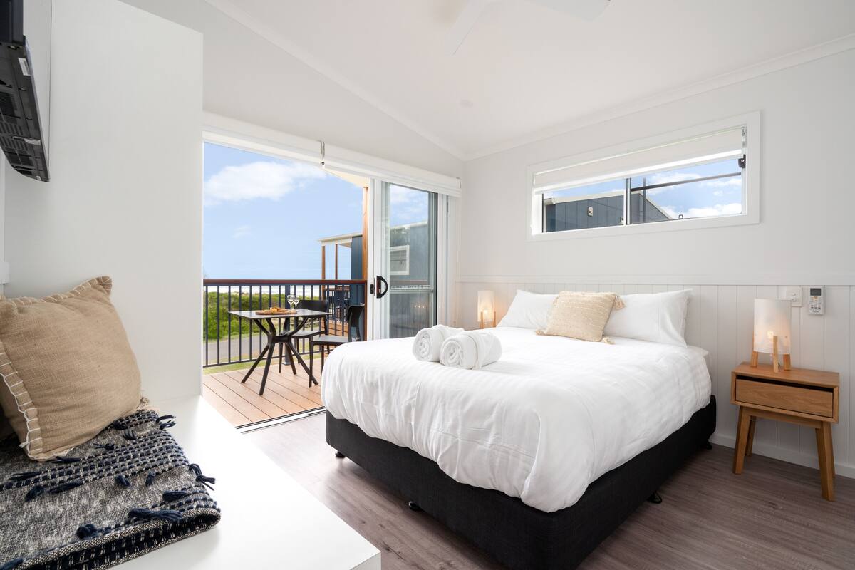 A bedroom with a large bed, a small table, and a balcony with a view.