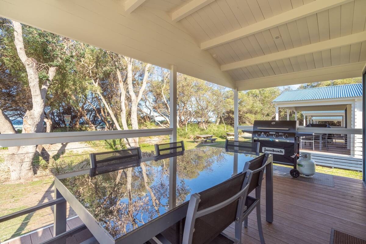 An outdoor patio with a glass table and chairs, a barbecue grill, and a view of trees and a house.