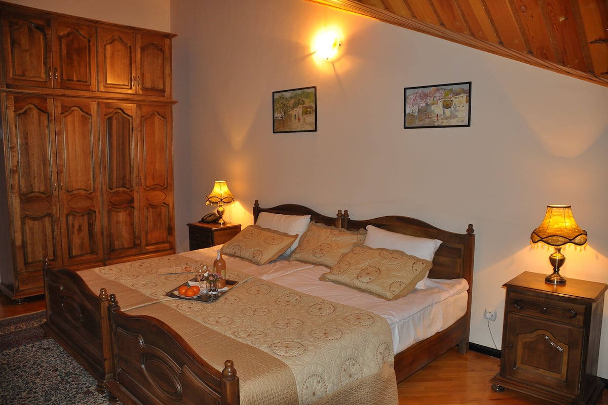 A bedroom with a wooden bed, two bedside tables with lamps, a wooden wardrobe, and two framed pictures on the wall.
