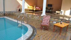 En swimmingpool med flisebelagte kanter, en træstol, et bord med en flaske og en seng i baggrunden.