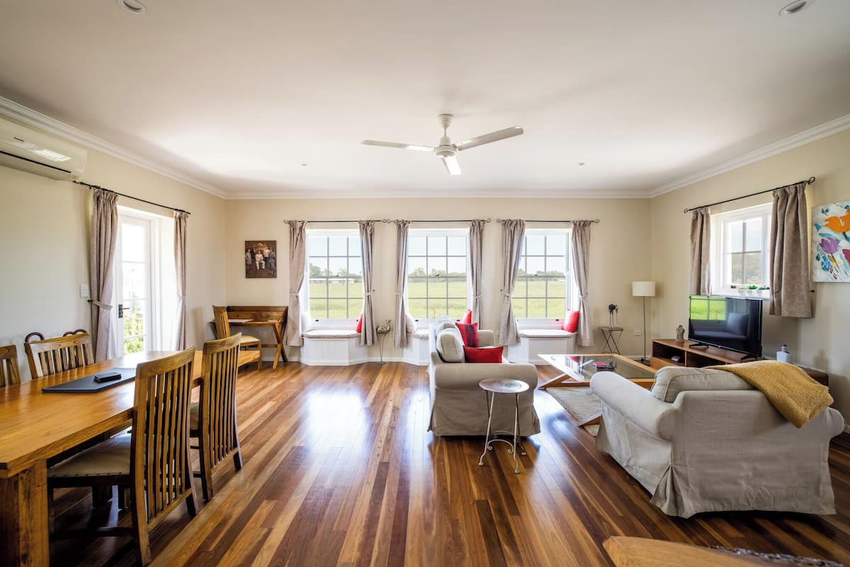 Un salón amplio con suelo de madera, un comedor con mesa y sillas de madera, un sofá, un televisor y ventanas grandes con cortinas.