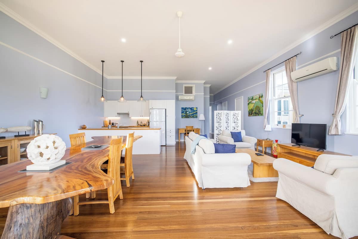 Un salón espacioso con una mesa de comedor de madera, sofás blancos y una cocina al fondo.