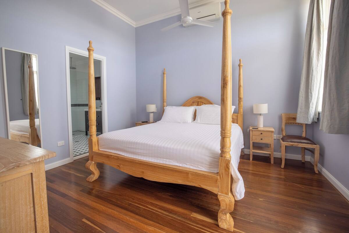 Una cama con dosel en una habitación con suelo de madera, ventilador de techo y mesita de noche con lámpara.