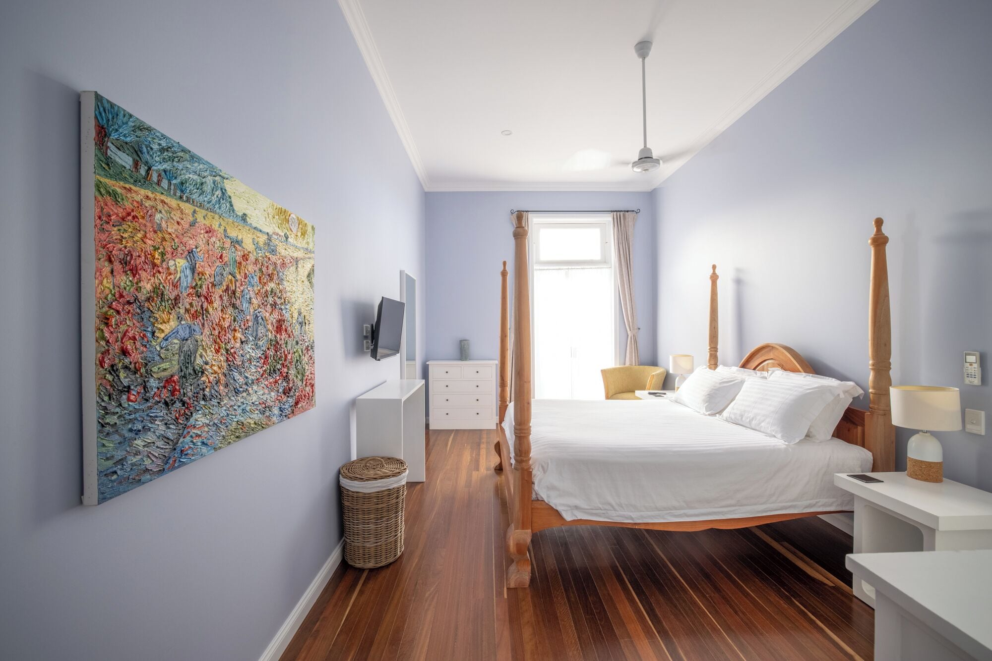 Un dormitorio con un cuadro grande, una cama con ropa blanca, un cabecero de madera, una mesita de noche con una lámpara y una pequeña cesta de mimbre.