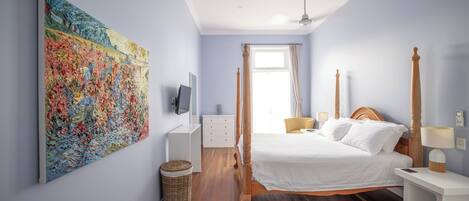 Un dormitorio con un cuadro grande, una cama con ropa blanca, un cabecero de madera, una mesita de noche con una lámpara y una pequeña cesta de mimbre.