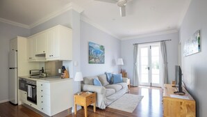 Une cuisine moderne avec des armoires blanches, un four encastré et un grille-pain. On y trouve une table à manger en bois, une lampe et un téléviseur. Le salon est doté d’un canapé beige, d’une table basse et d’un tableau accroché au mur.