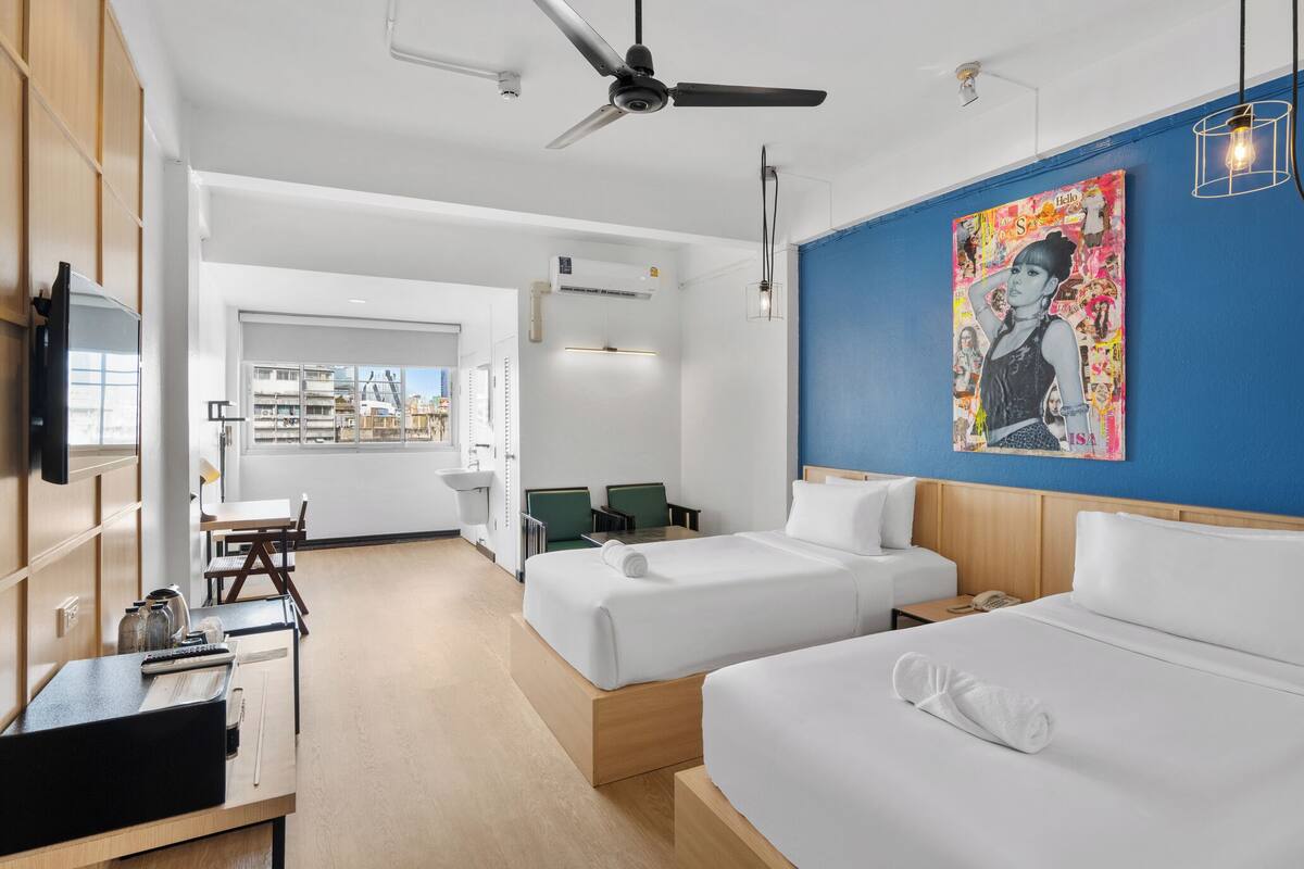 Ein modernes Hotelzimmer mit zwei Betten, einem Schreibtisch, einem Stuhl, einem Fernseher und einem großen Fenster mit Blick auf die Stadt.