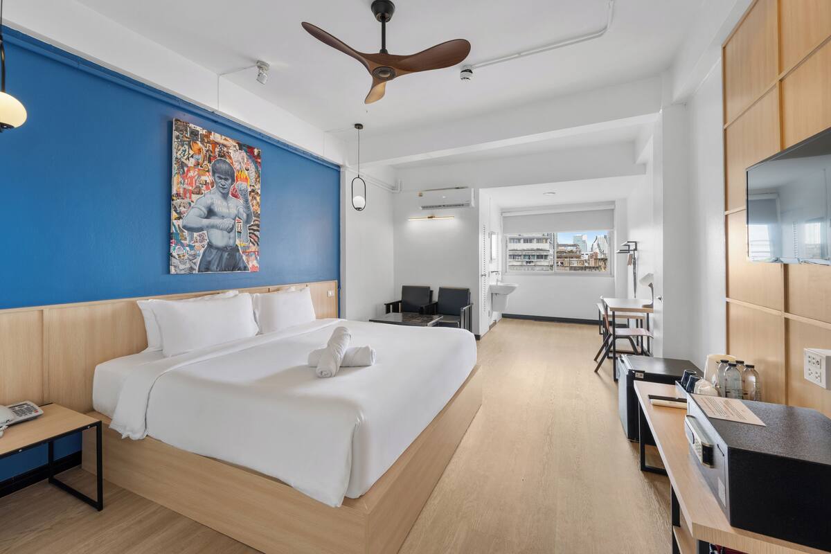 Ein modernes Hotelzimmer mit einem großen Bett, einem Deckenventilator, einem Schreibtisch mit Computer und Blick auf die Stadt durch das Fenster.