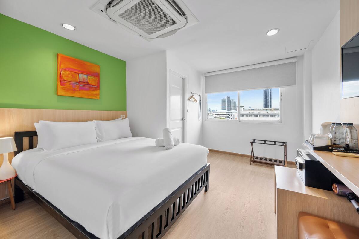 Ein Hotelzimmer mit einem großen Bett, einem Schreibtisch mit Kaffeemaschine, einem Fenster mit Blick auf die Stadt und einer Wand in Grüntönen.